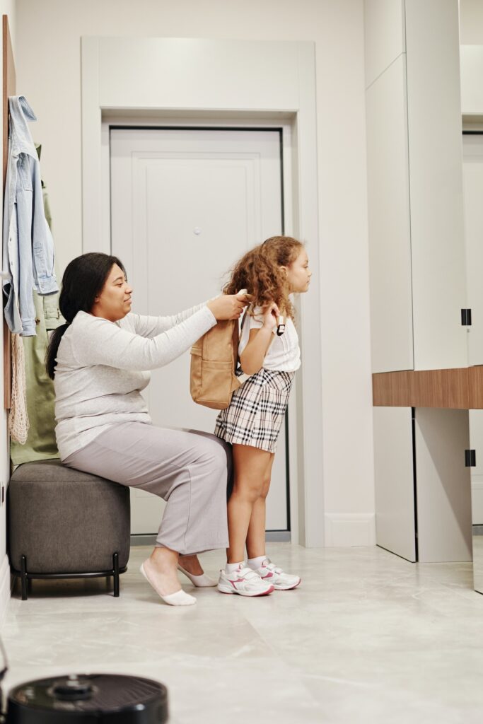 Mom helping her daughter prepare for school by putting her backpack on her back and supporting her as part of the ADHD therapy.