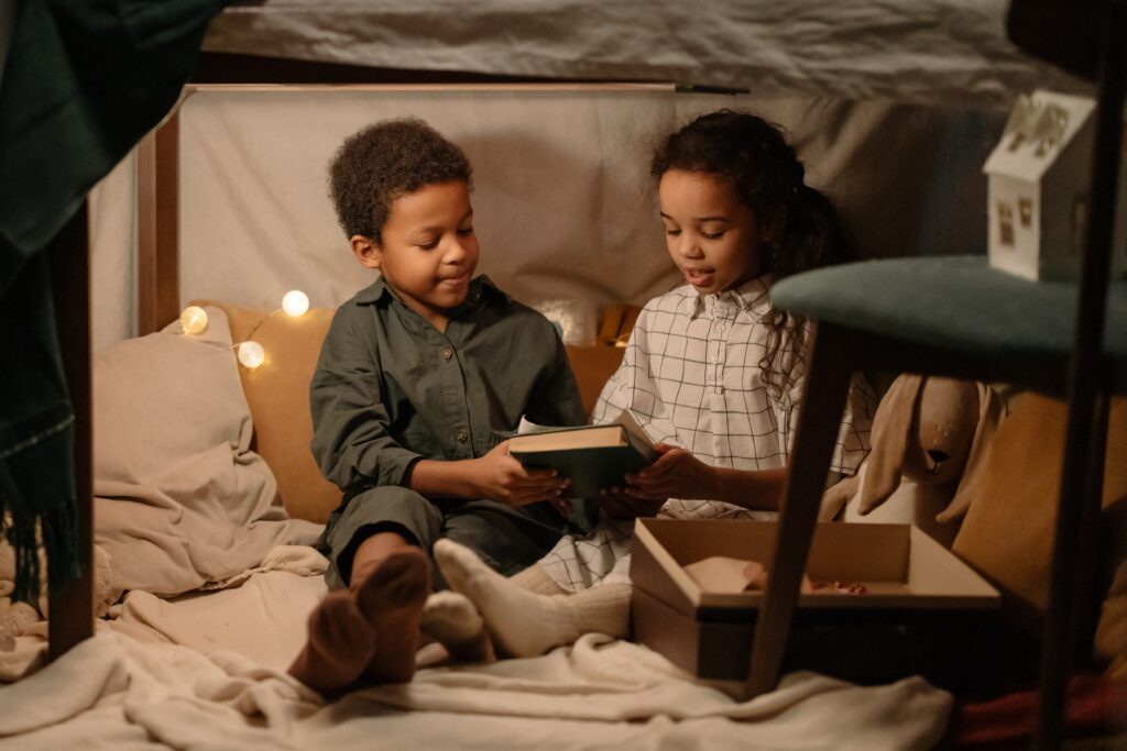 A little girl recommends a book to her friend while they are sitting in a home-improvised tent with blankets and pillows.